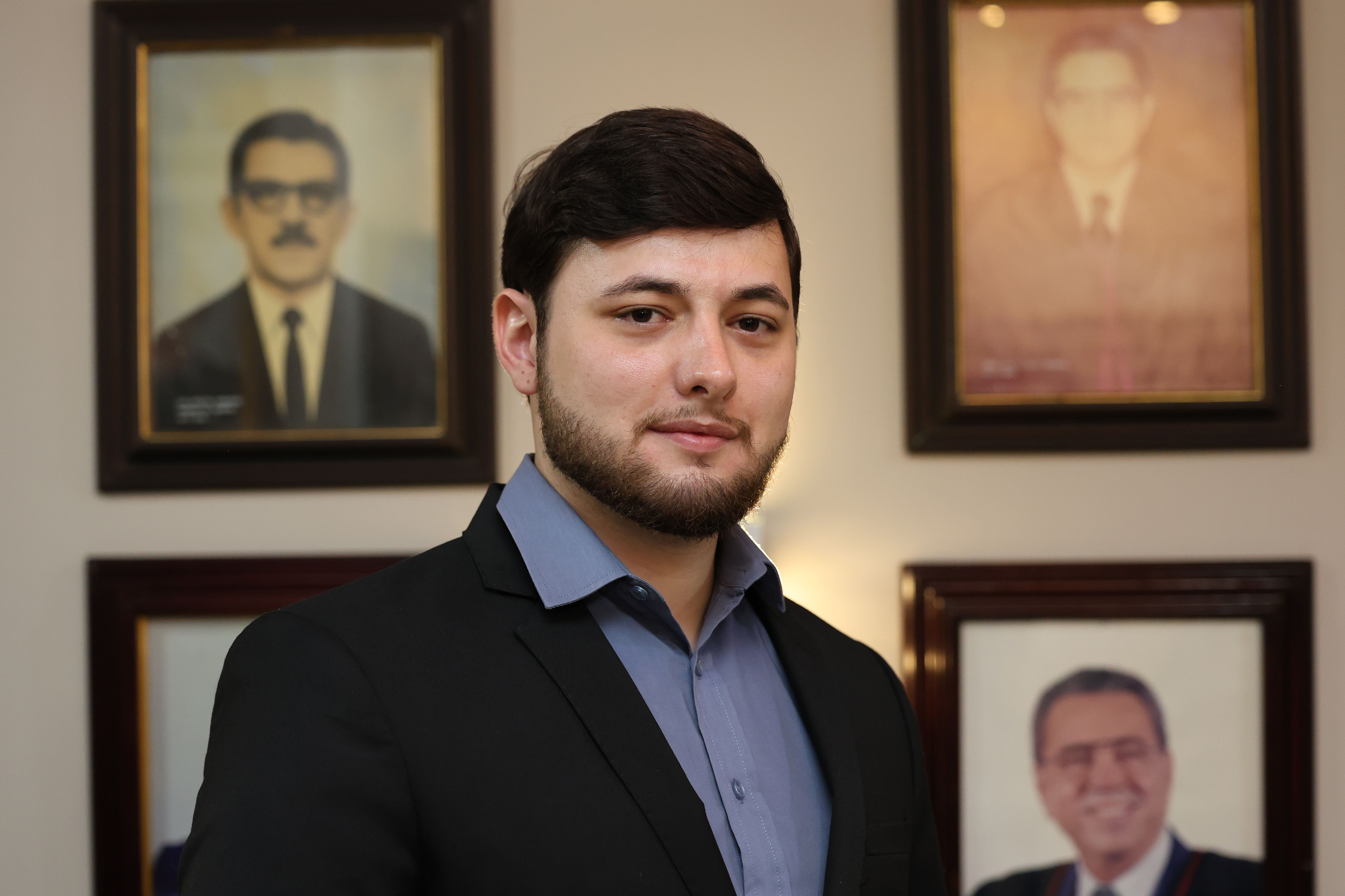 Foto 2 - Paulo José Amorim  Um homem jovem, com barba, cabelo castanho escuro e terno preto sobre camisa azul, está em pé. Ele olha para a frente em um ambiente interno. Atrás dele, há quatro retratos emoldurados de pessoas na parede.