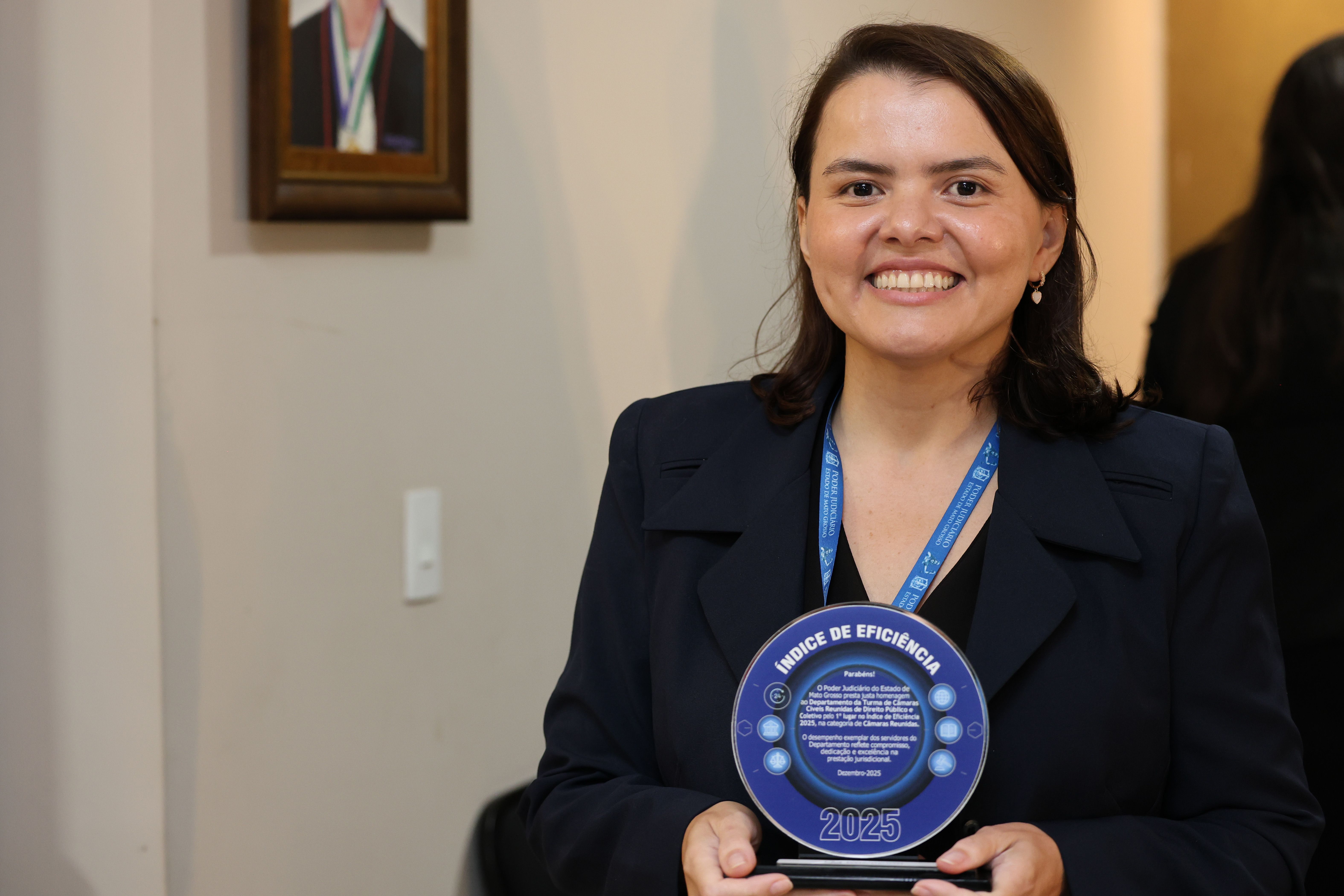 Foto 3 - Nayara Iraidy - Uma mulher de cabelos longos, morena, com blusa azul-escura, segura um prêmio redondo e azul com a inscrição 
