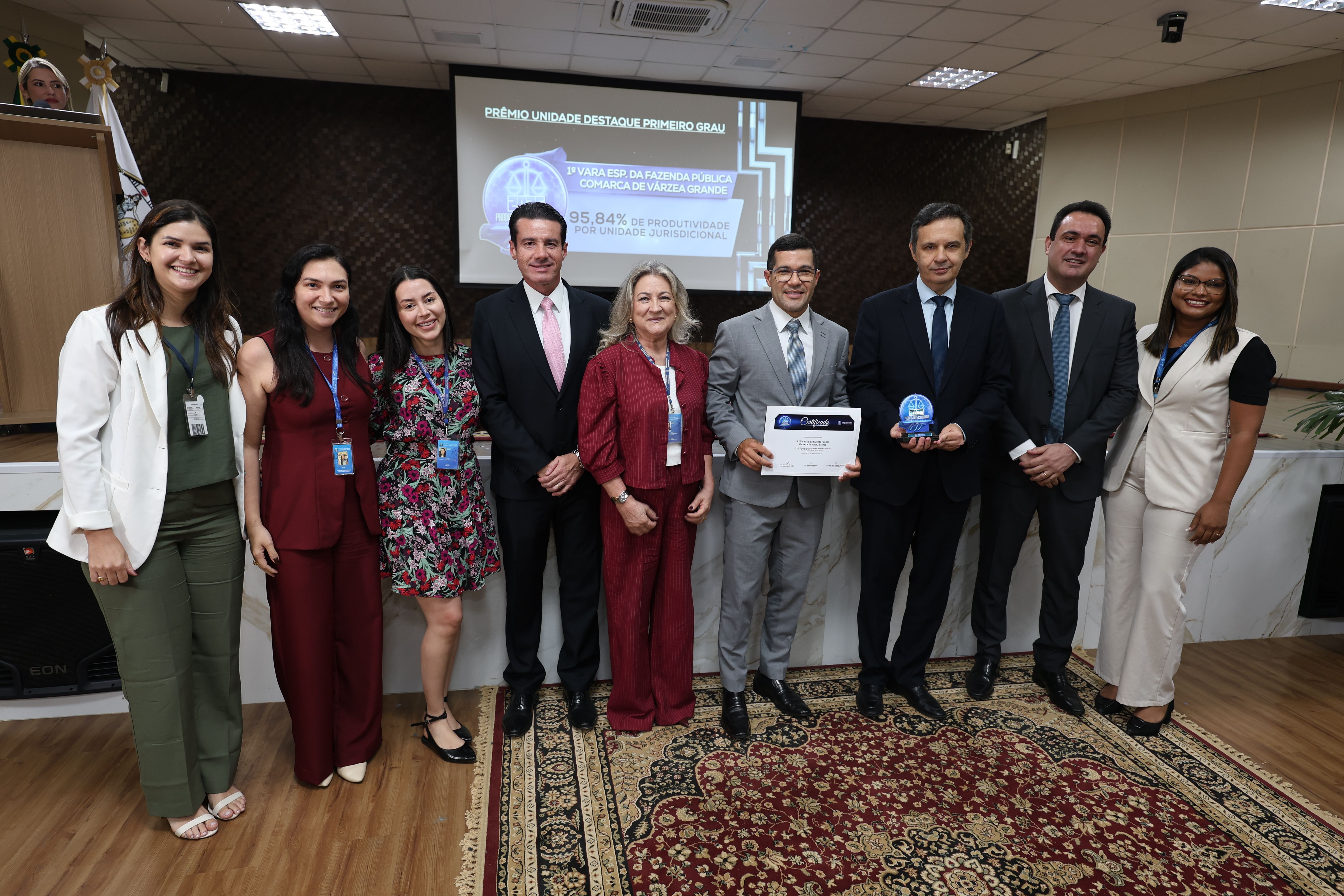 Foto 4 - juiz Ramon e servidora Divania - Um grupo de oito pessoas (quatro homens e cinco mulheres) está em pé em um auditório, em frente a um telão com o prêmio 